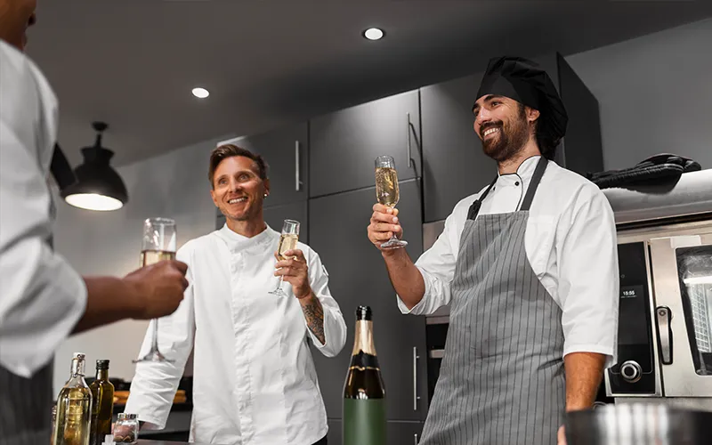 chef-celebra-con-equipo-buen-trabajo Chef y cocineros brindando por el éxito del restaurante, la buena comunicación y el fortalecimiento del trabajo en equipo en la cocina profesional.